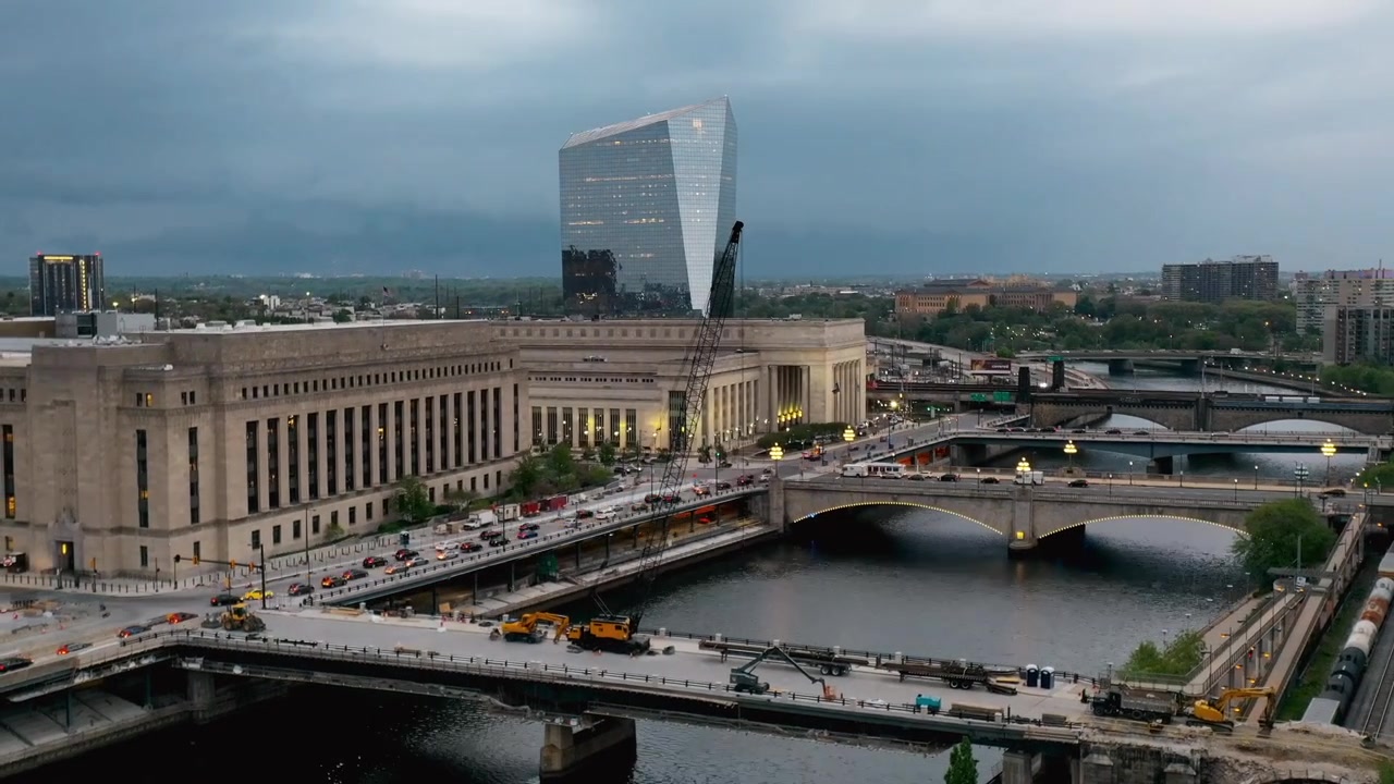 Luftfoto af Philadelphia, der viser trafik, der bevæger sig over broer over Schuylkill-floden, med den historiske 30th Street Station og den moderne Cira Centre-bygning, der dominerer skyline ved skumringstid.