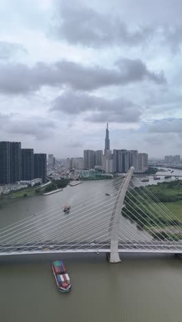 Container-boats-on-Saigon-River,-Ho-Chi-Minh-City,-with-high-aerial-view-of-Ba-Son-suspension-Bridge-with-landmark-building-in-vertical-format-as-boat-passes-under