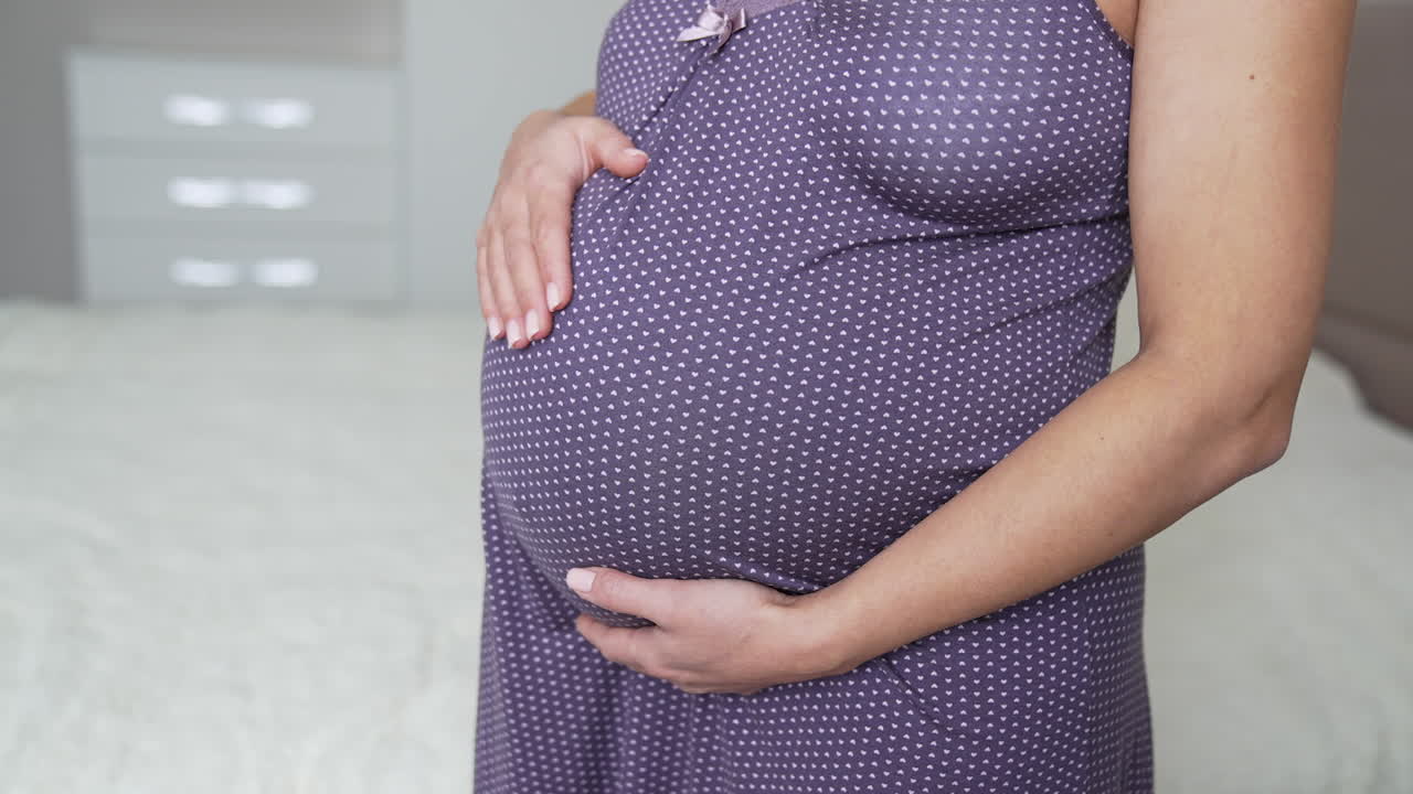 Future mother in nightdress is in the light bedroom. She caresses her belly with one hand. Circle movements of hands around pregnant tummy.