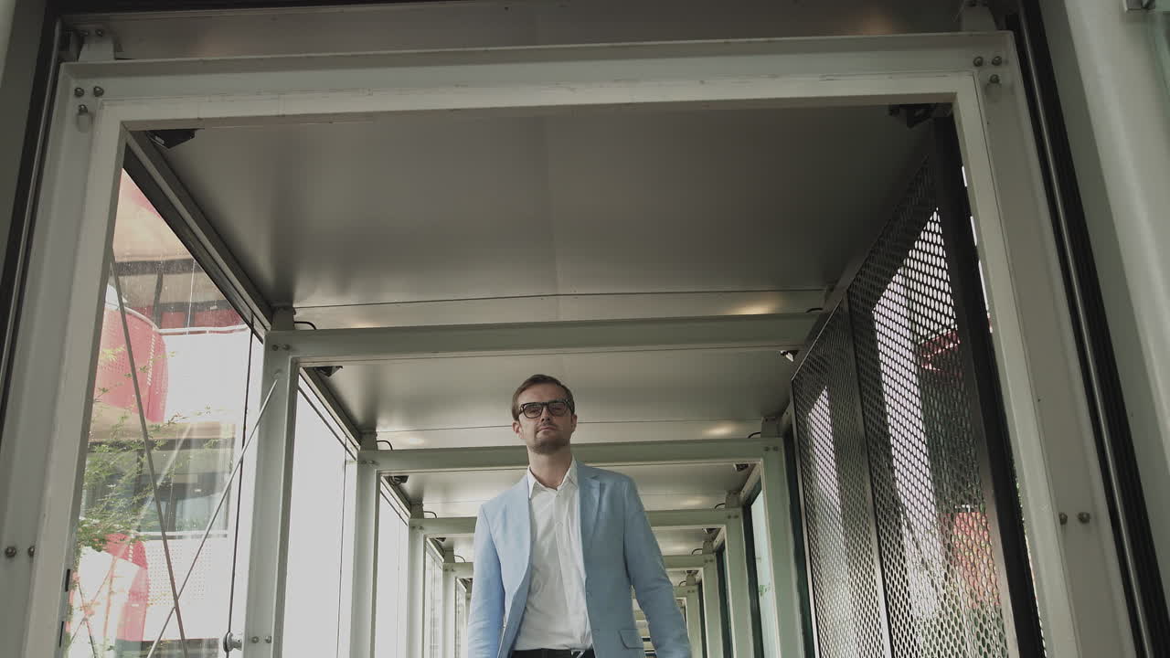 Un giovane uomo con una giacca e degli occhiali alla moda si avvicina sicuro di sé alla fotocamera in un moderno corridoio di vetro