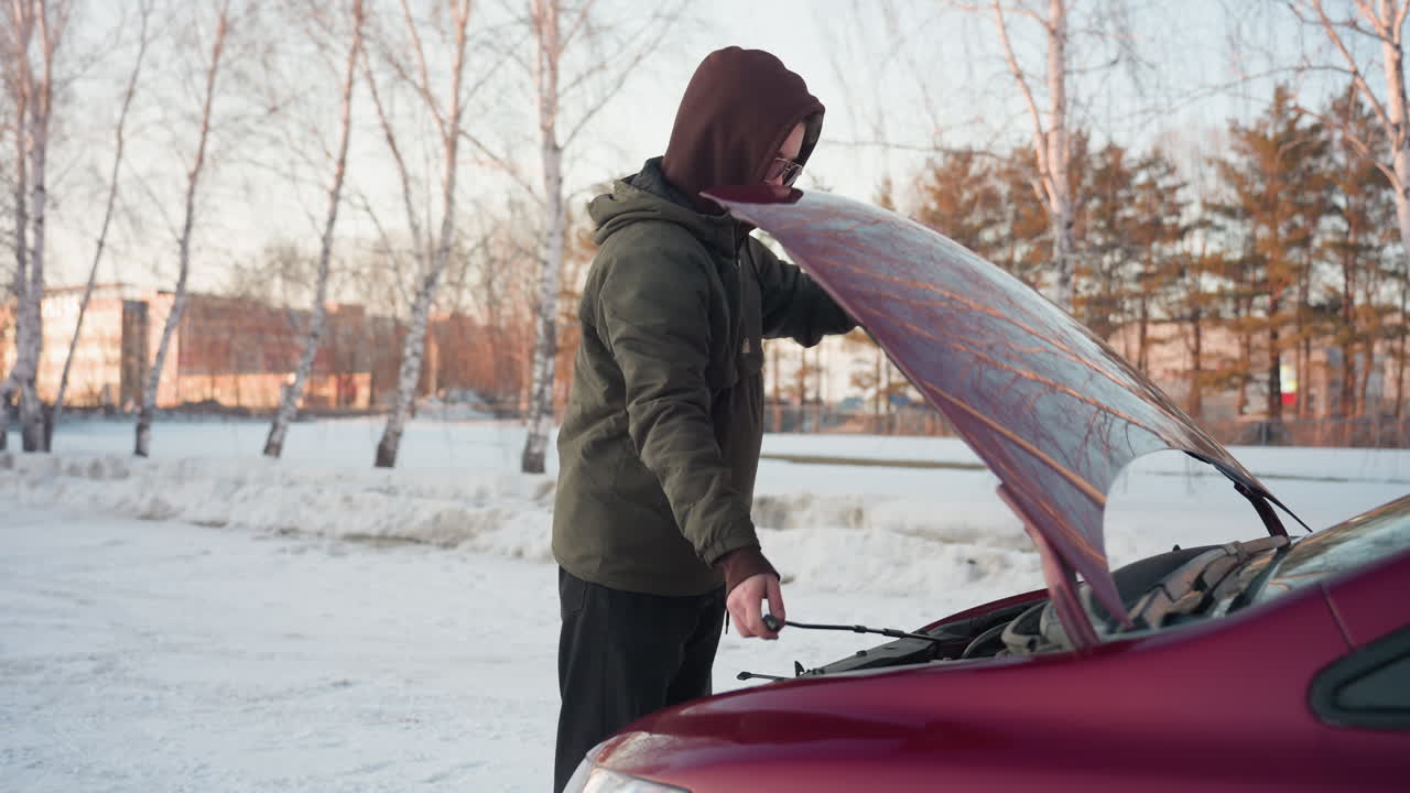 Mechanic wearing hooded winter jacket closes raised car bonnet after inspecting engine outdoors in snowy environment with bare trees and distant building in background