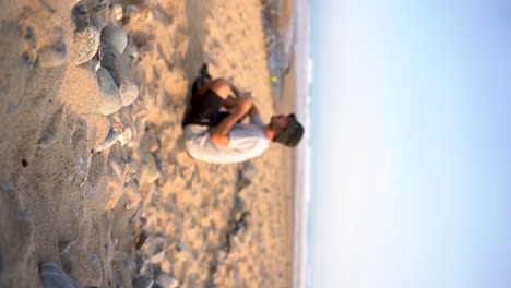 Joven-Vertical-Sentado-En-La-Playa-Bebiendo-Café-Matutino-Bajo-El-Cálido-Sol-Del-Amanecer