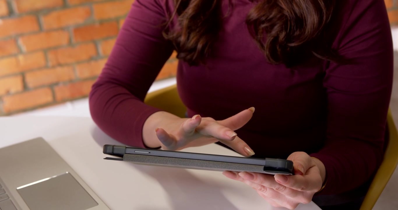 Female in maroon top holding tablet at round table, steadying left, swiping right, laptop left