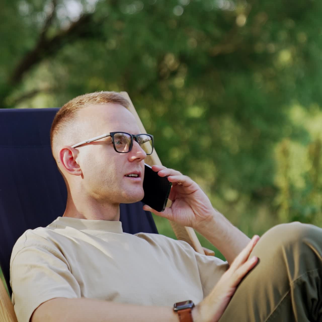 Confident male businessman sitting in the chair outdoors. Man talks on the phone at backdrop of nature
