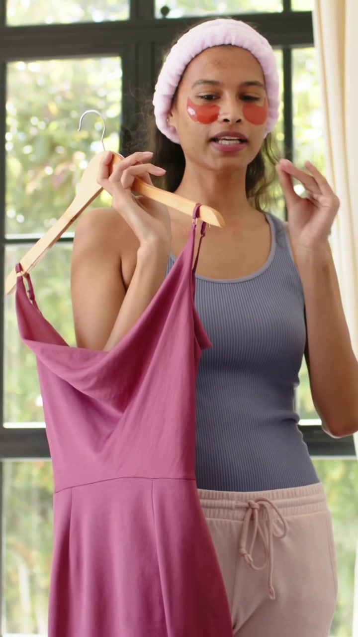 Vídeo vertical: Mulher de regata azul puxando cabide, segurando vestido magenta, verificando o caimento em casa.