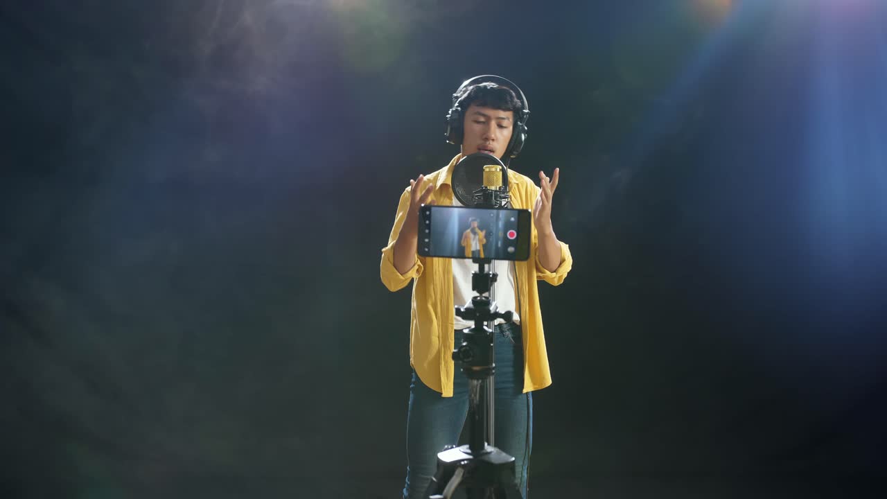 joven asiático con auriculares cantando en un micrófono de condensador mientras graba video con un teléfono inteligente en el fondo blanco de humo negro