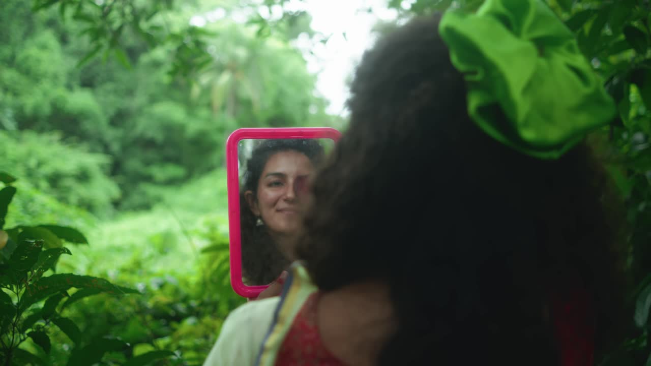 Primo piano di una donna in sari vista da dietro, che tiene uno specchio in una vegetazione lussureggiante. Il suo riflesso la mostra sorridente e che si copre un occhio con un petalo di fiore prima di girarsi verso la telecamera.