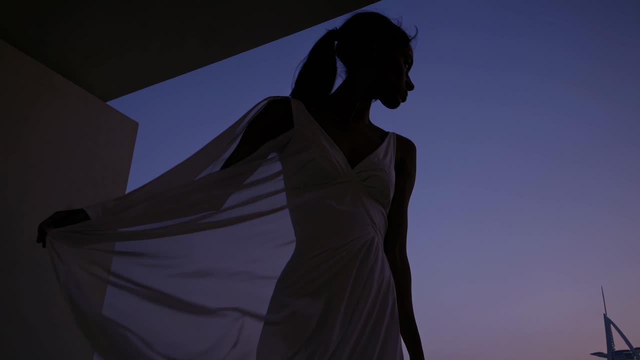 Silhouetted woman gracefully posing on a balcony at sunset, with the iconic Burj Al Arab hotel rising in the background, embracing the tranquil twilight ambiance of Dubai