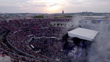 Drone-over-the-Arena-of-Nimes-at-sunset-and-stage-smoke,-people-are-waiting-for-the-stromae-concert