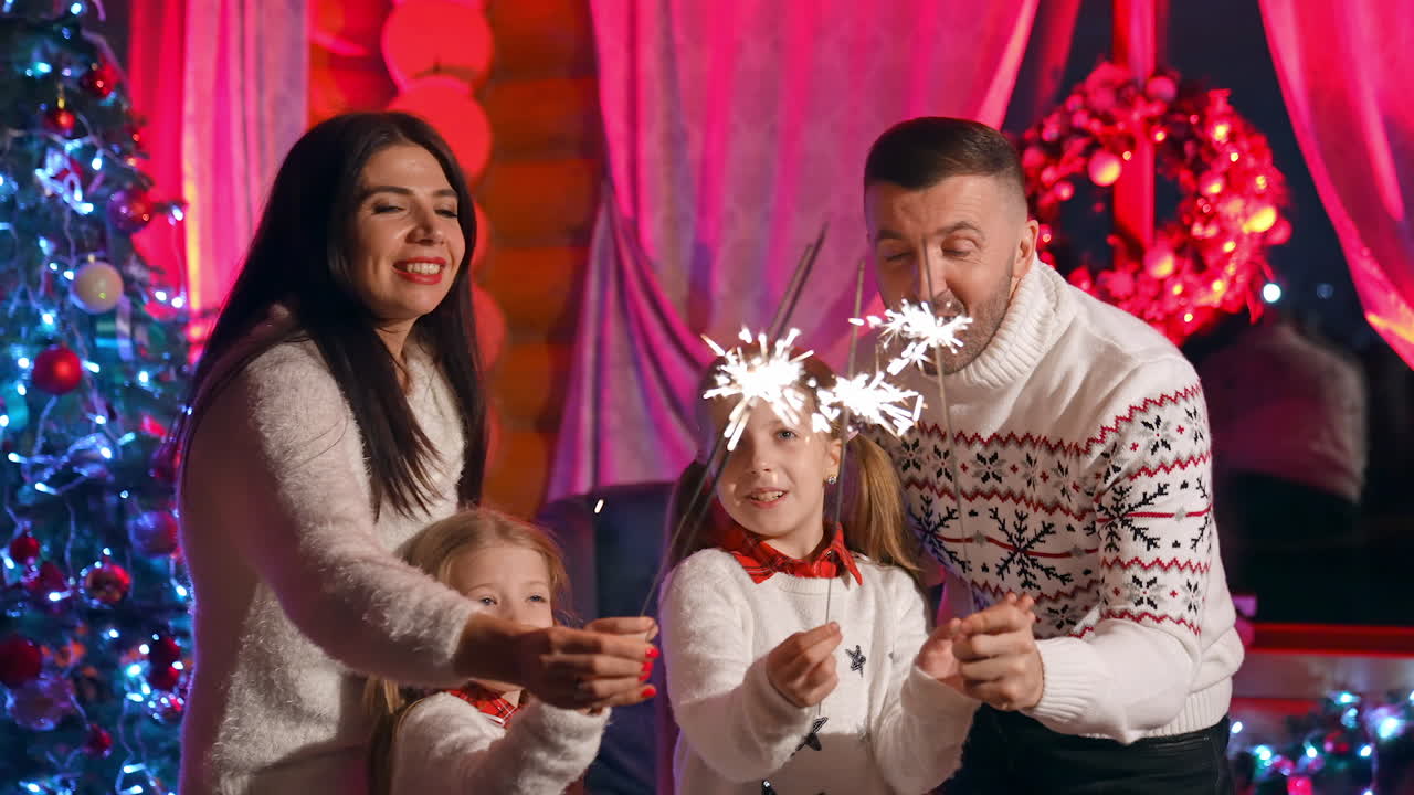 Happy family celebrate New Year at home. Parents and their daughters in warm white sweaters holding sparkler lights. All family members happy together at Christmas.