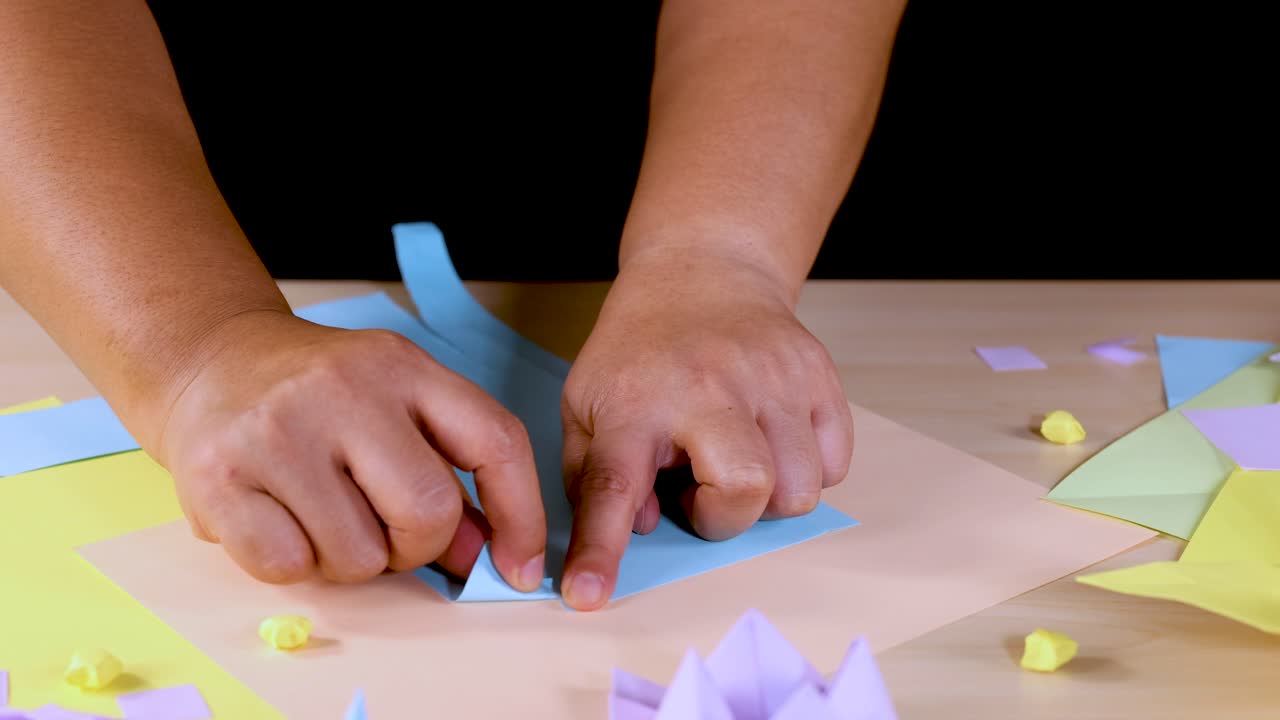 Hands tear blue paper on a craft table with colorful origami, under bright studio lighting
