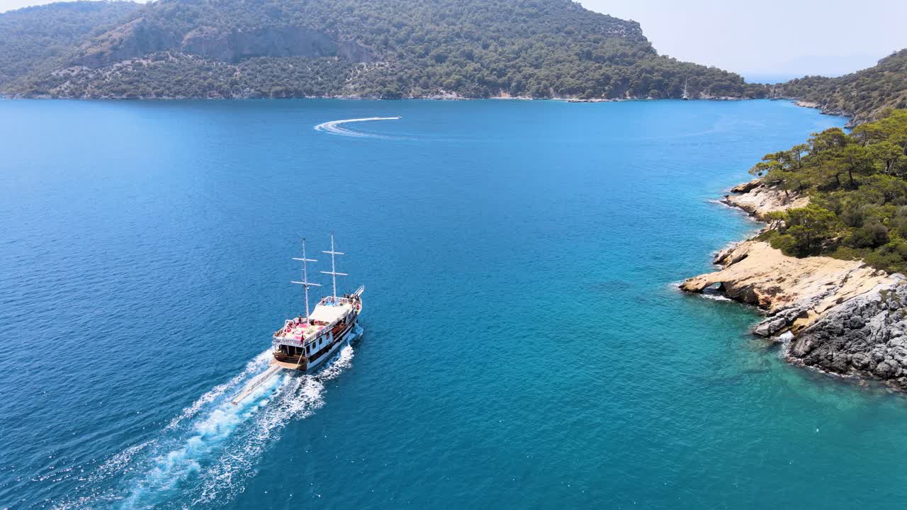 Drone fly over a bay in the Mediterranean over a yacht sailing between islands covered in green trees under a blue sky