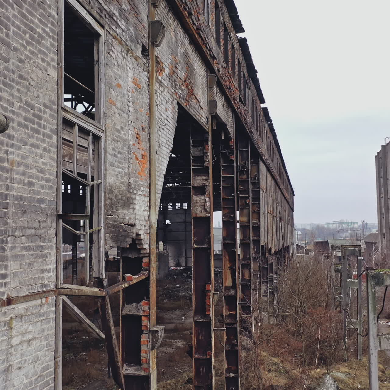 Former plant building with damaged brick walls and metal structures covered with moss on the roof and overgrowths inside and outside. Aerial view.