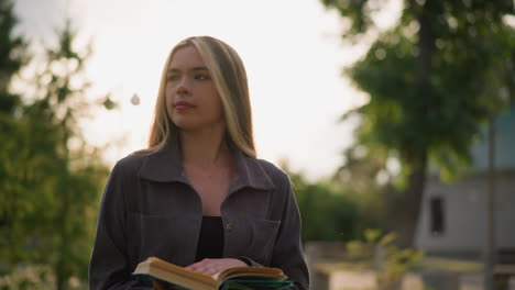 lady in grey clothing walking on grassy field while reading a book as sunlight creates bright flashes around her, looking thoughtful with a warm smile, with a blurred sunlit background