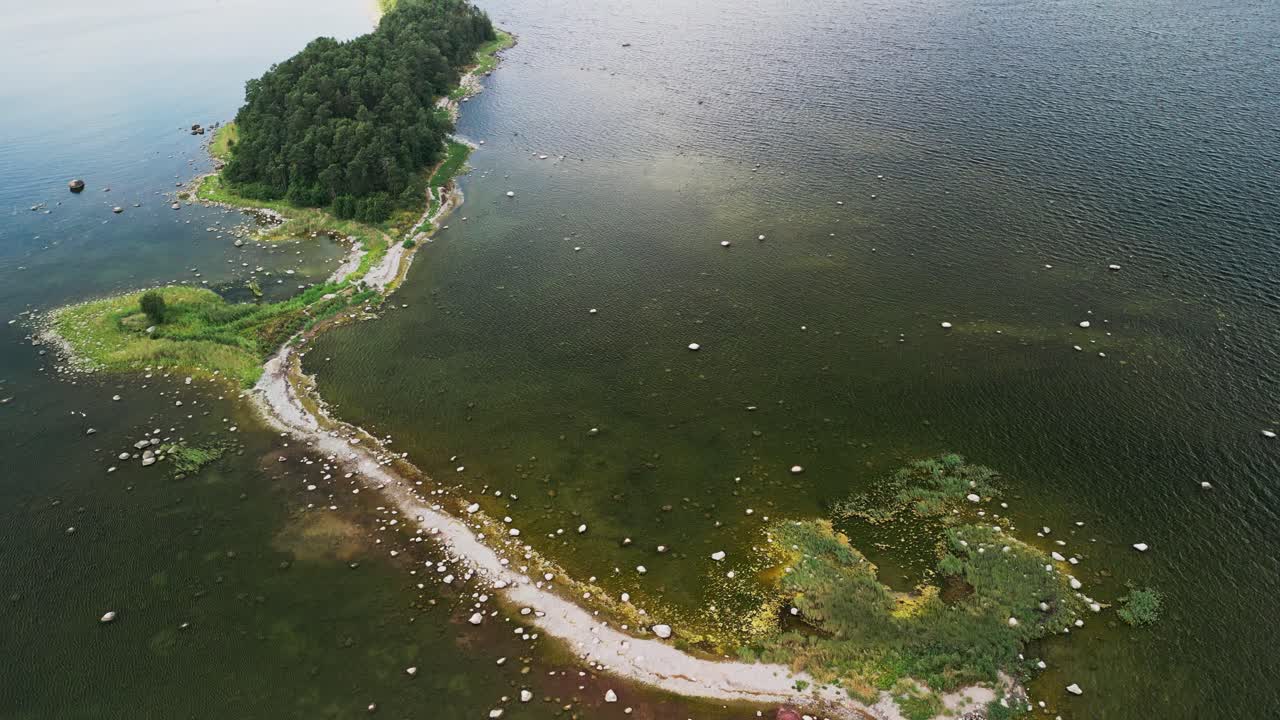 Captivating drone view of Estonia’s rugged coast highlights a forest island linked by a narrow land path, walkable at low tide, with rocky shoreline formations and calm Baltic waters