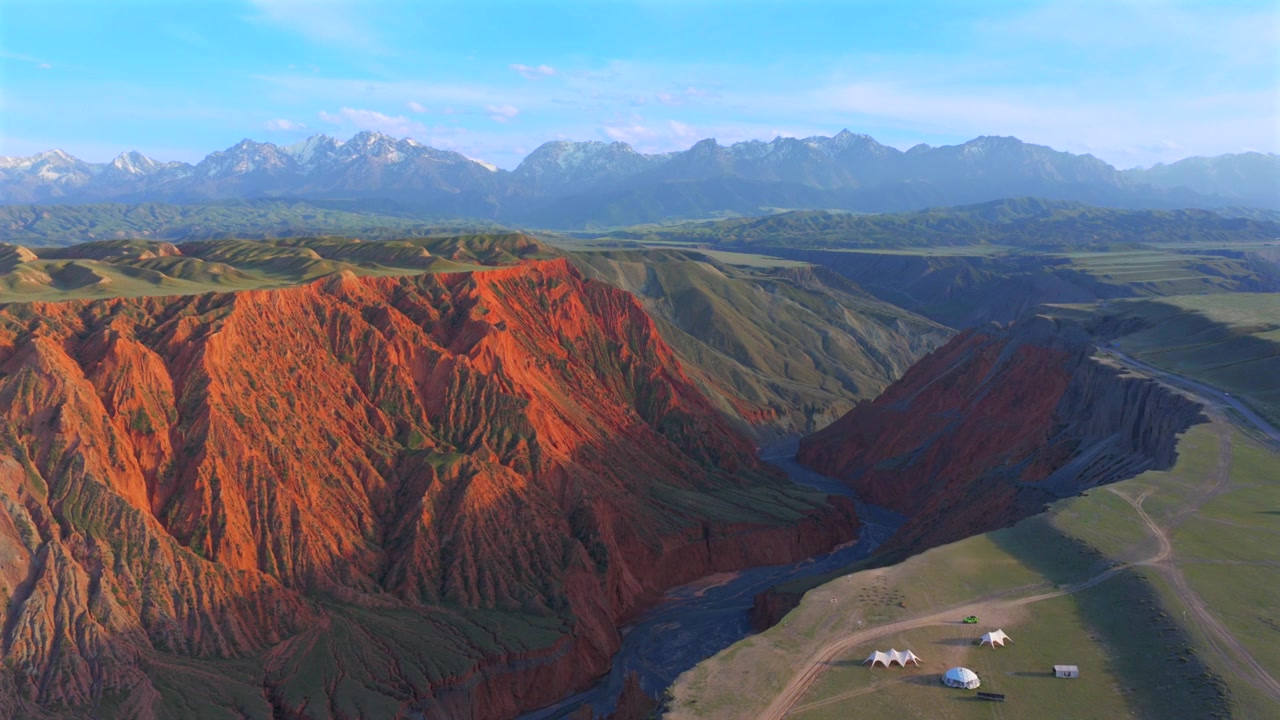 Sincan (Xinjiang) seyahat noktasında su erozyonuyla oluşmuş geniş kanyon arazisi, katmanlı kırmızı kaya uçurumları ve karla kaplı Tanrı Dağları (Tianshan) zirvelerinin drone görüntüsü