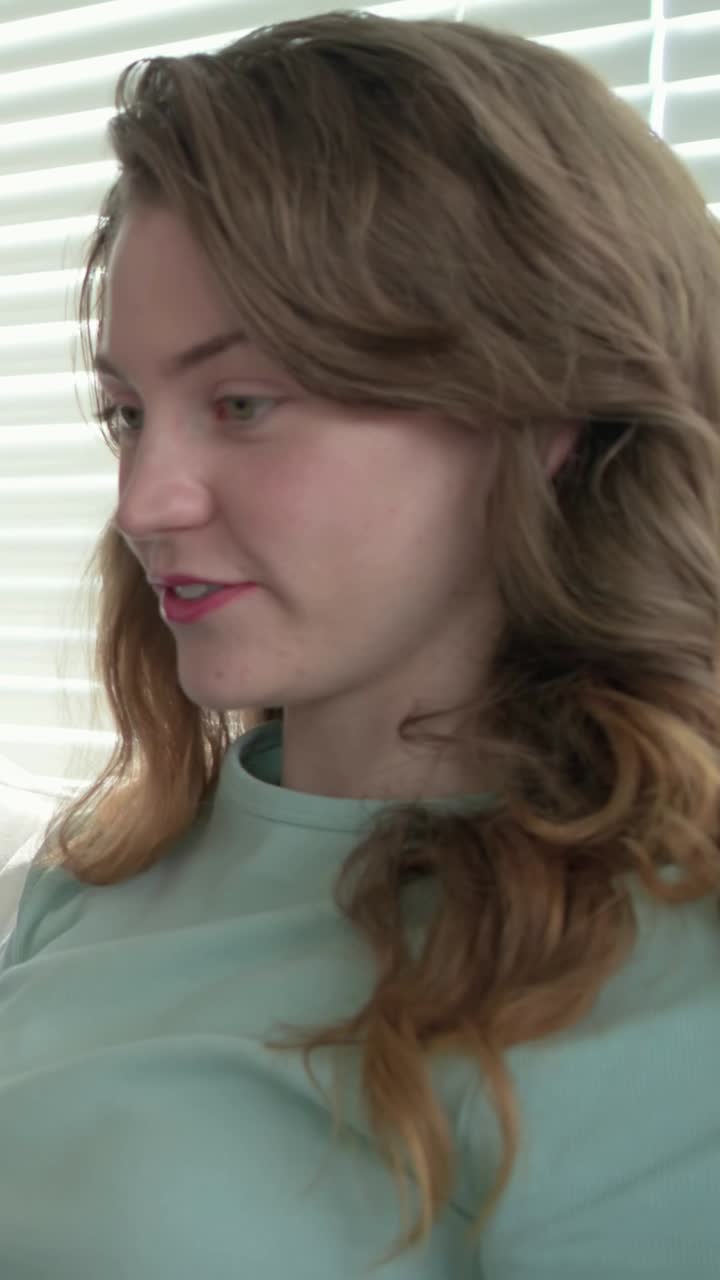 Vertical video: Turning woman in light green tee glancing left as blue arm showing on sofa, blinds
