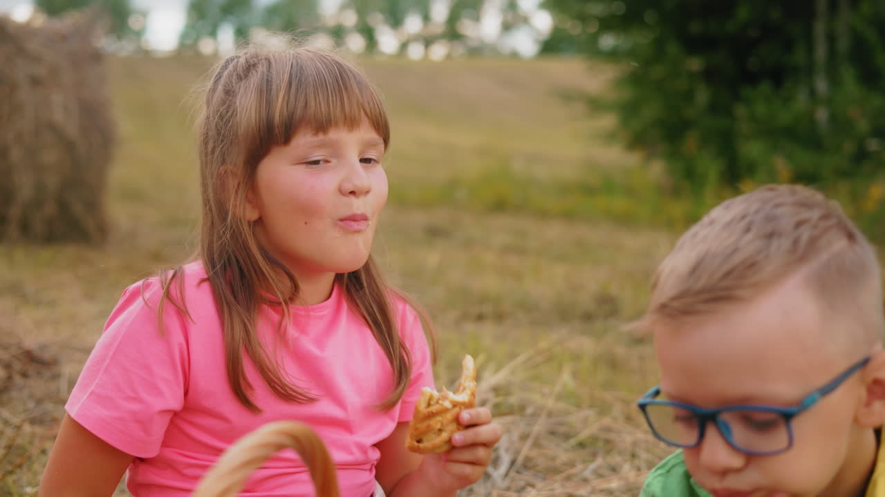 Una bambina si gusta i suoi spuntini all'aperto con una vista parziale della testa di un bambino seduto lì vicino, entrambi si godono i dolci in un sereno campo aperto circondato dalla natura e da un paesaggio tranquillo.