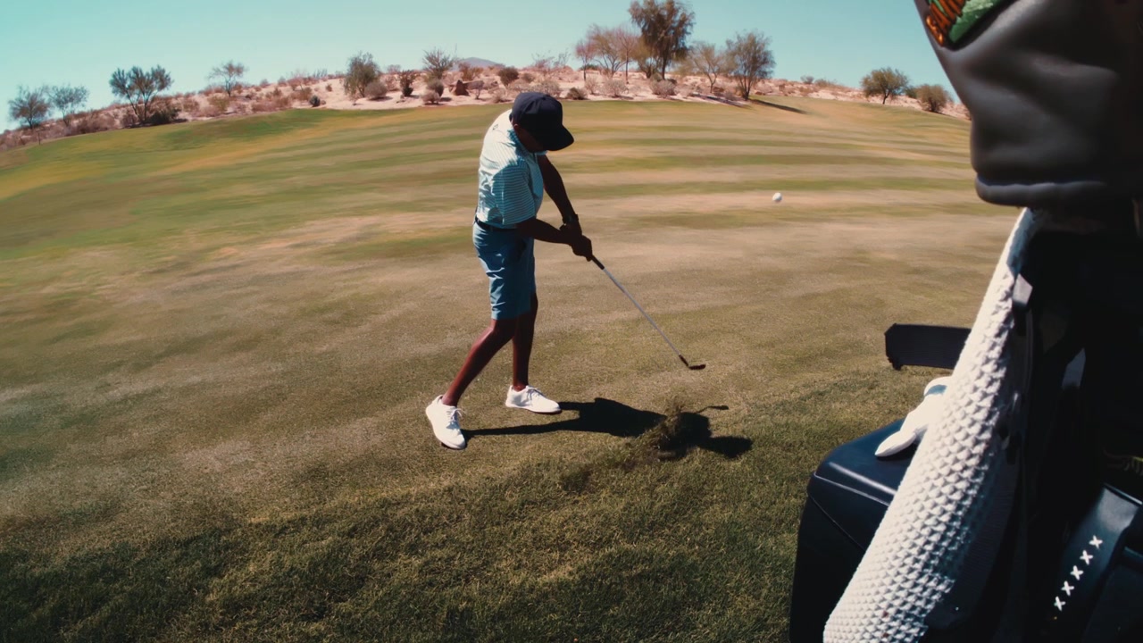 Un golfista colpisce un colpo con il ferro dal primo taglio di rough in ultra slow motion, ripreso in 4K a 800fps oltre una sacca da golf con colline desertiche aride sullo sfondo.