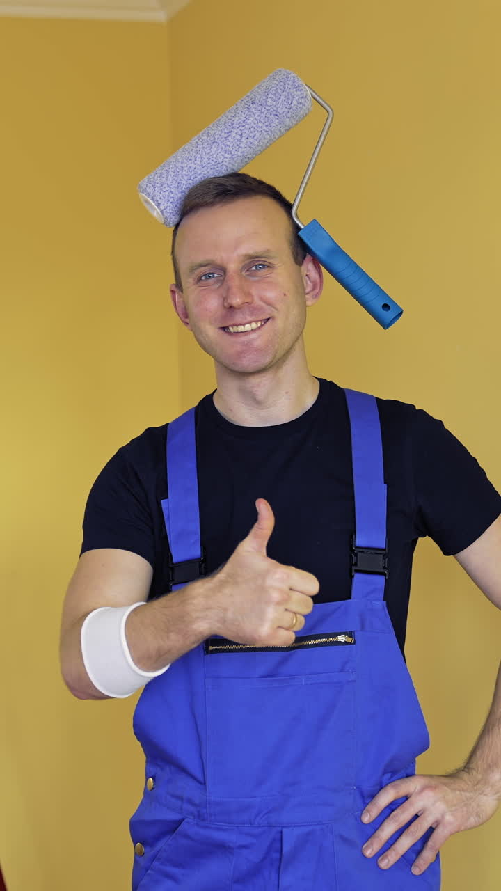 Portrait of a painter man indoors. Young smiling worker in overalls with a painter roller on his head showing yes sign with a thumb. Renovation services. Vertical video