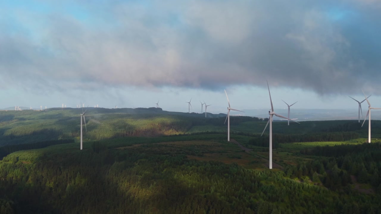 High Contrast Morning View of Large Onshore Wind Farm Spread Over Mountainous Terrain Landscape with Forest and Dramatic Sky.