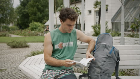 young man reading a booklet, sitting on a banch next to his grey backpack