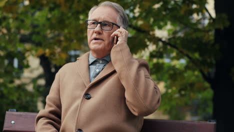 elderly man in glasses and coat sitting on the bench and talking on the phone in the park in autumn