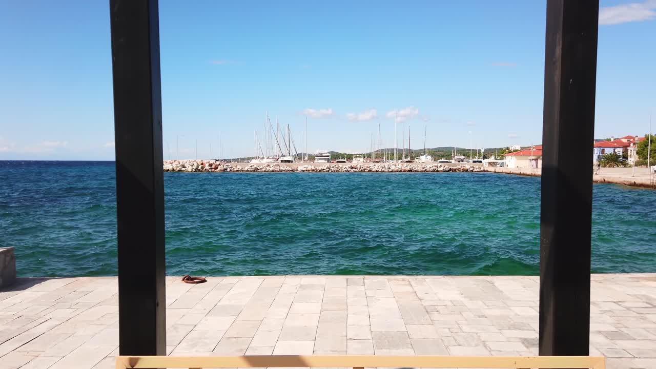 Blick auf einen Seehafen mit vertäuten Yachten, Ägäisches Meer, Blick von einem Pavillon auf einem Pier mit schwarzen Metallpfosten und einer Bank in Nikiti, Griechenland. Zeitlupe