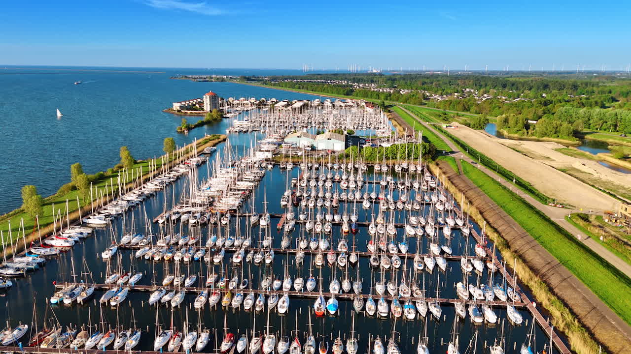 Flight over the multiple yachts at the berths in the marina of Lelystad, the Netherlands. Lush greenery at the waterfront at backdrop. Aerial view.