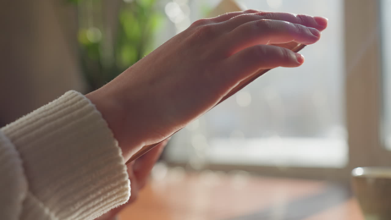 Close up of hand flipping page of open book in soft daylight near window, warm ambient light casting glow on paper, coffee mug in background on wooden table