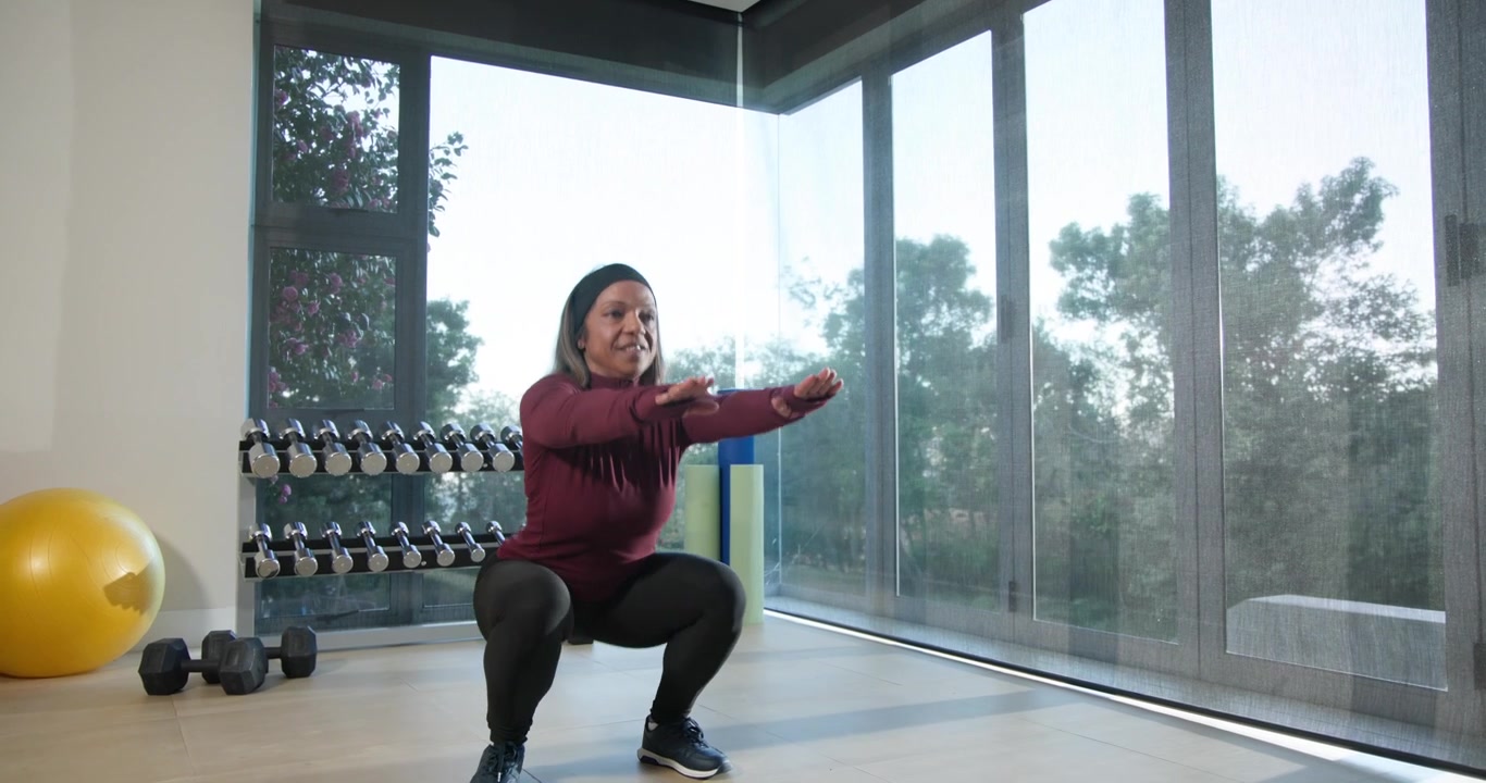 Donna afroamericana che fa squat seguendo una routine in una palestra domestica, con top bordeaux, leggings e pesi.