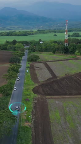 Aerial drone shot capturing a self-driving car linked to a tower on a countryside road. Overcast skies and lush greenery define the serene vertical video setting. Ideal for mobile use