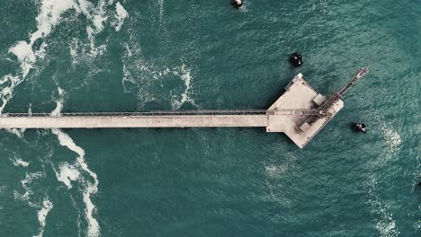 Aerial of gravel loader jetty extending into blue ocean, waves crashing around structure, top down ascend