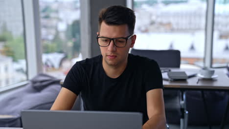 young caucasian man puts on glasses to work at computer. freelancer typing on his laptop sitting in the spacious room. blurred backdrop.