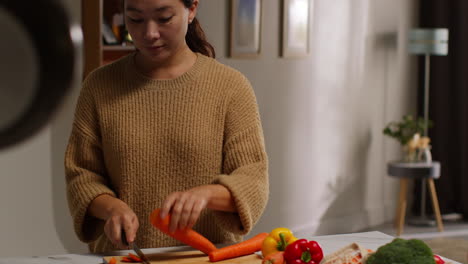 woman at home in kitchen preparing healthy fresh vegetables for vegetarian or vegan meal chopping carrots on board