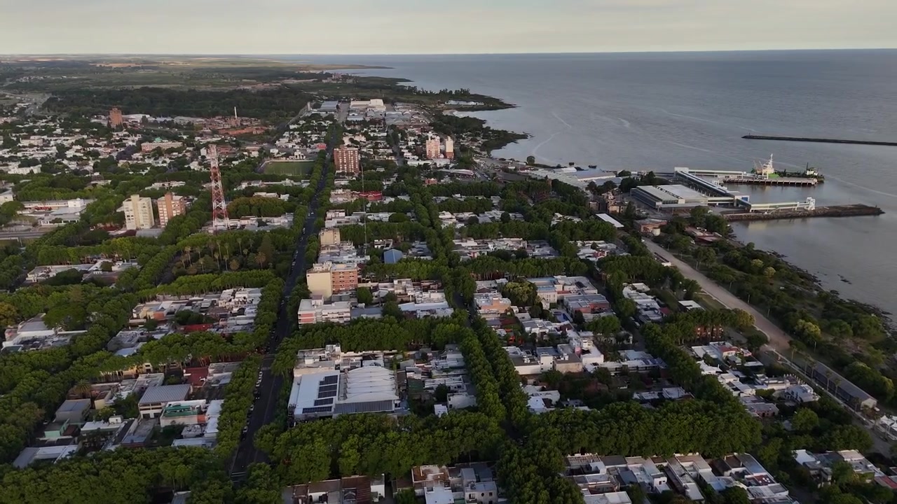 Skyline del lungomare cittadino con strade alberate, grattacieli, porto fluviale con nave ormeggiata, torre radio a Colonia del Sacramento, Uruguay, calda luce del tramonto. Ripresa aerea panoramica con drone