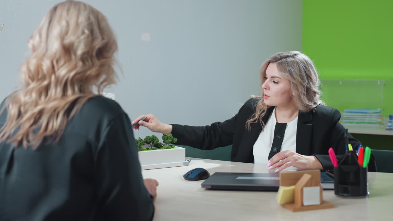 Saleswoman explaining modern residential building miniature to client during detailed discussion at office table, pointing with pencil while describing project layout