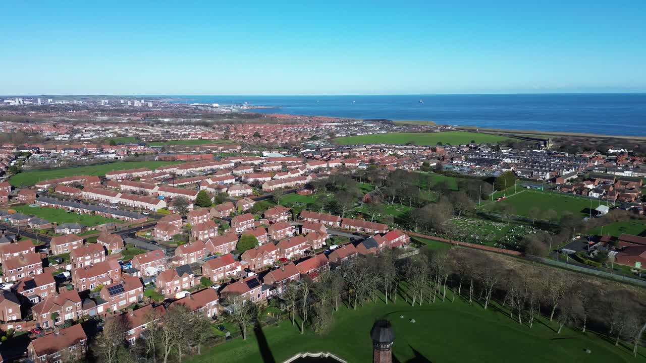 Drohnenluftaufnahme über dem Dorf Ryhope, Sunderland, die rechts in Richtung Nordseeküste abbiegt. Ein klarer blauer Himmel offenbart Vorstadtdächer, Straßen, Grünflächen und den fernen Meereshorizont.