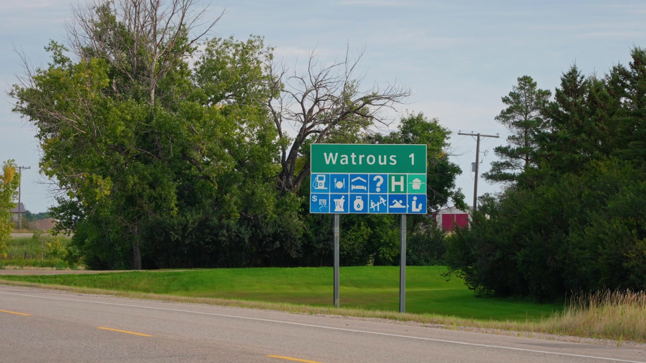 Static telephoto shot of a highway sign outside Watrous, Saskatchewan, with trees in the background, road in the foreground and a car driving across the frame