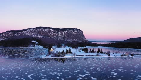 Orbitando-Alrededor-De-Una-Península-En-Un-Lago-Congelado-En-La-Base-Del-Acantilado-De-Una-Montaña-Aislada-Al-Amanecer-Con-Patrones-De-Líneas-Y-Círculos-En-El-Hielo