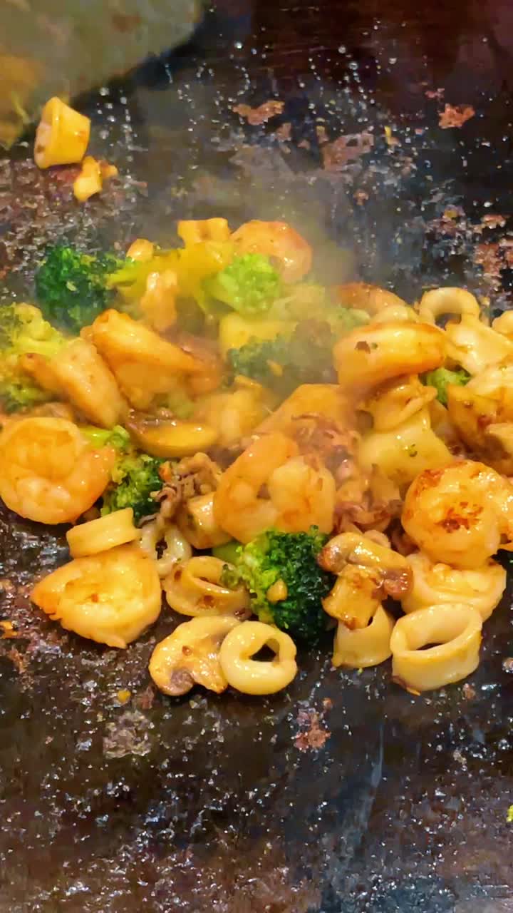 Chef che cucina frutti di mare e verdure su piastra a cottura piana. Mani che usano spatole di metallo per cucinare gamberi, anelli di calamaro e broccoli su una piastra professionale in acciaio inossidabile.