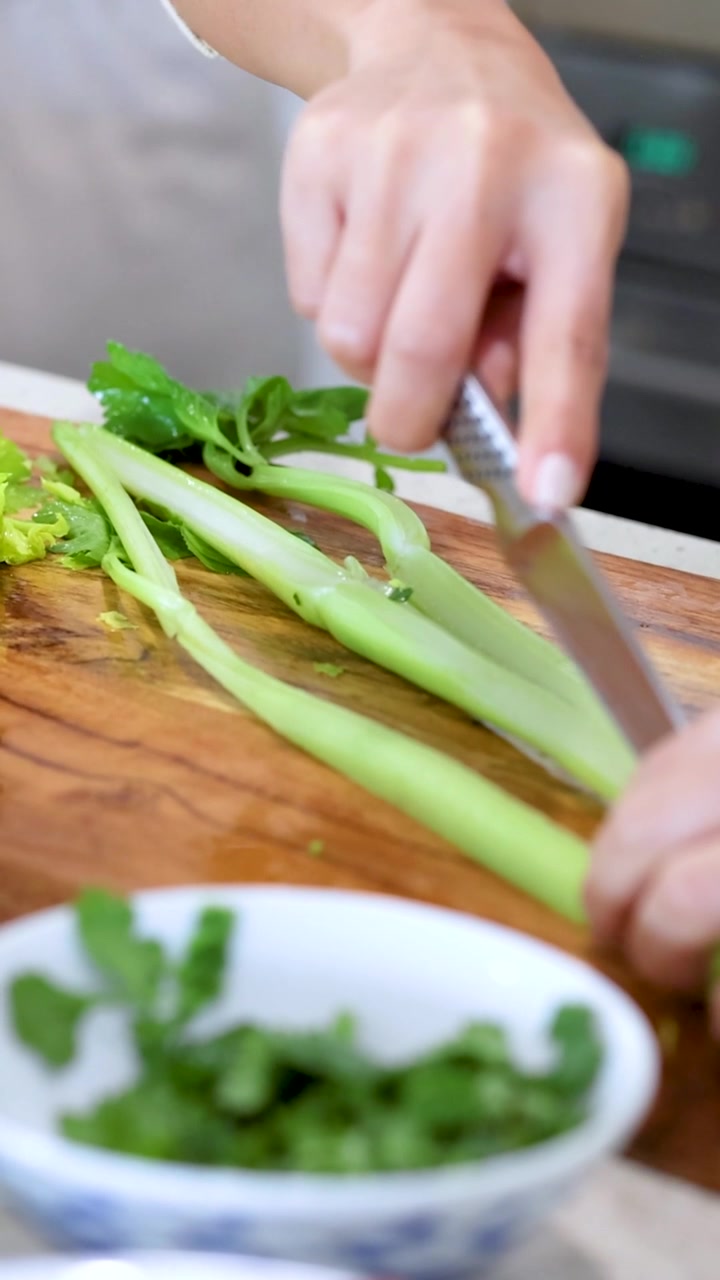 A close-up vertical shot of hands using a knife to slice celery on wood.