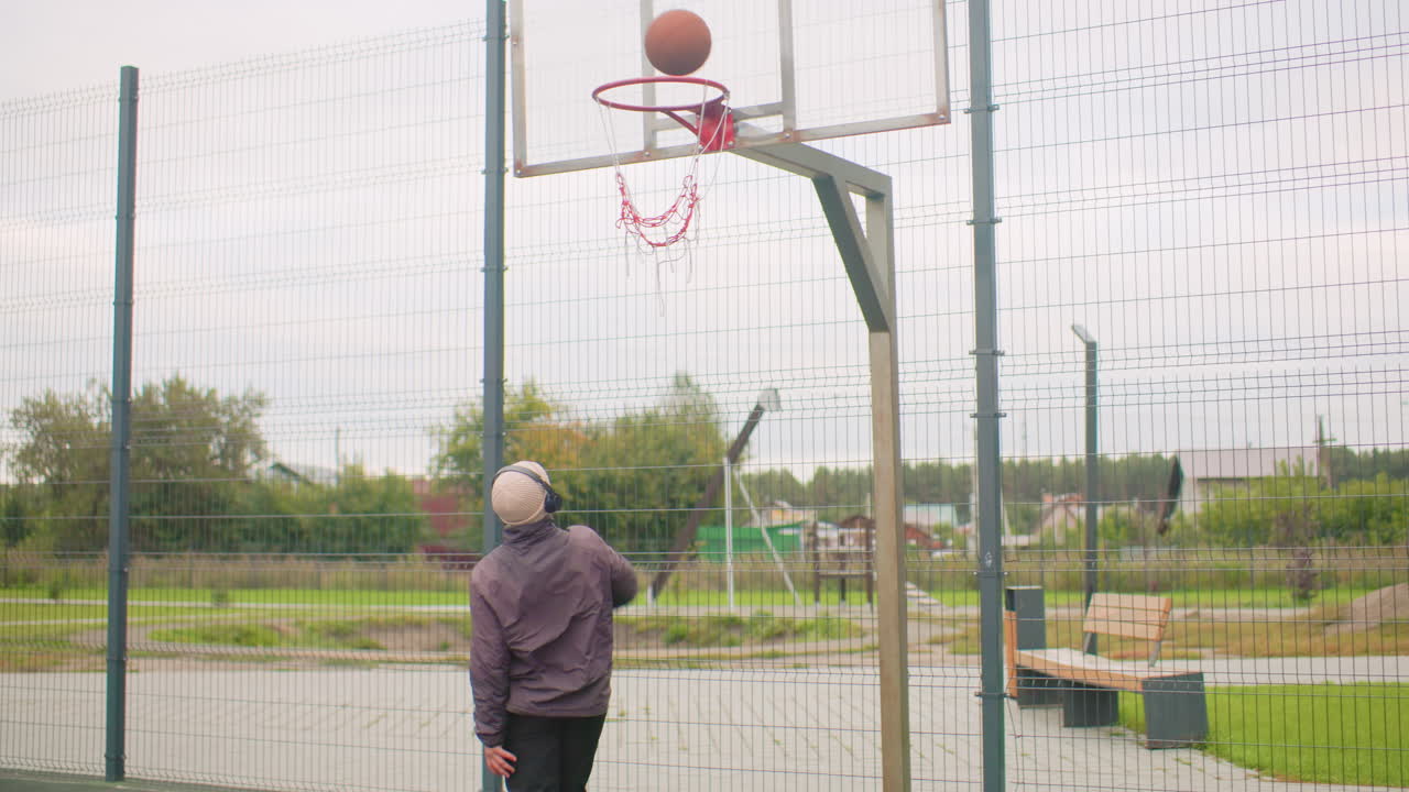 Giovane che si allena a basket all'aperto in un campo urbano recintato vicino a condomini, che indossa un berretto e cuffie, palleggia e si dirige verso il canestro, tira a canestro e guarda la palla colpire il cerchio, solitario
