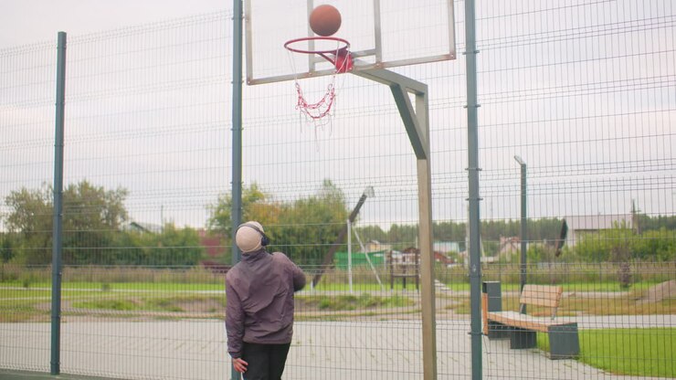 Giovane che si allena a basket all'aperto in un campo urbano recintato vicino a condomini, che indossa un berretto e cuffie, palleggia e si dirige verso il canestro, tira a canestro e guarda la palla colpire il cerchio, solitario