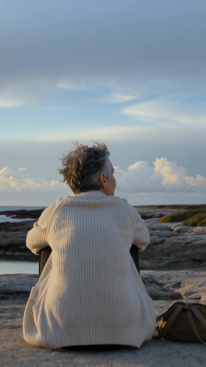 Vertical video: Sitting senior in cream knit scanning as clouds shift on rocky shore, canvas bag