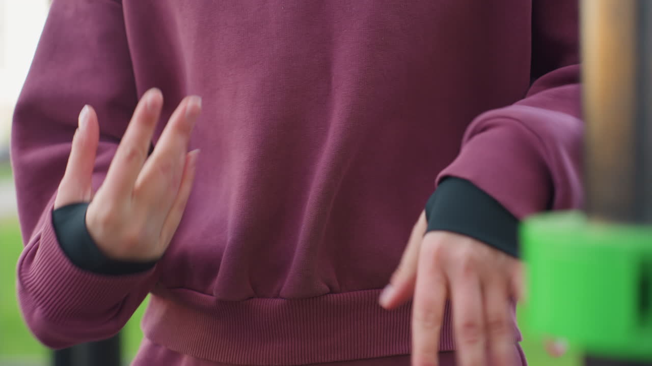 Stylish jogger hoodie performs gentle hand stretch by pressing palms together and flexing fingers during calm relaxation phase before outdoor workout under open sky at urban fitness park
