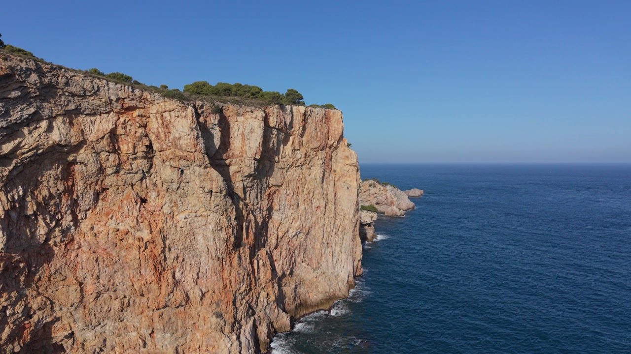 Rising drone footage of high steep cliffs with trees on the top against clear blue sky with deep blue sea and gently lapping waves at their base on bright sunny day