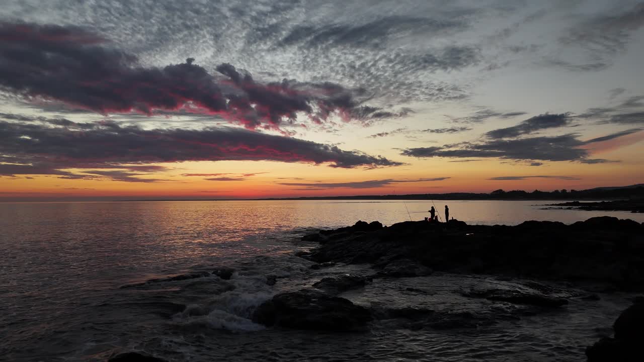 At Playa Verde, Uruguay, the drone orbits two fishermen silhouetted on rocks against vibrant post-sunset sky. Dramatic clouds and sea glow in magical twilight light
