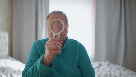 elderly woman in green jacket holding magnifying glass, focusing on object through lens, sitting on bed in bright room, with serious expression and natural light streaming through window