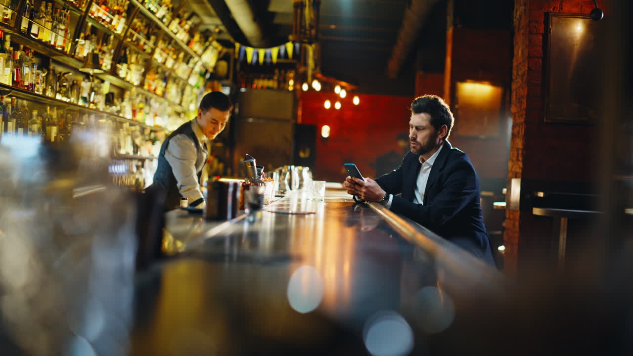 Un cliente rilassato al bar manda messaggi con lo smartphone seduto al bancone. Un ragazzo beve cognac